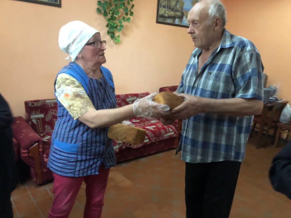 Elderly woman wearing a headscarf and gloves hands a loaf of bread to an older man inside a modest room with red patterned furniture and a warm-coloured wall.