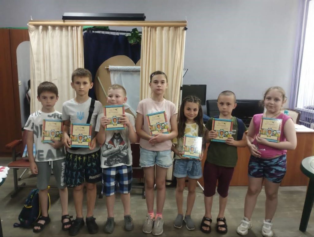 Seven children standing in a row indoors, each holding a copy of the Jesus Storybook Bible, smiling or posing for the camera during a summer camp