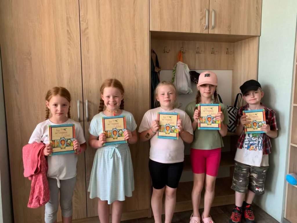 Five young children standing indoors in front of wooden cabinets, each holding a copy of the Jesus Storybook Bible and smiling holding copies of the Jesus Storybook Bible, part of a summer camp