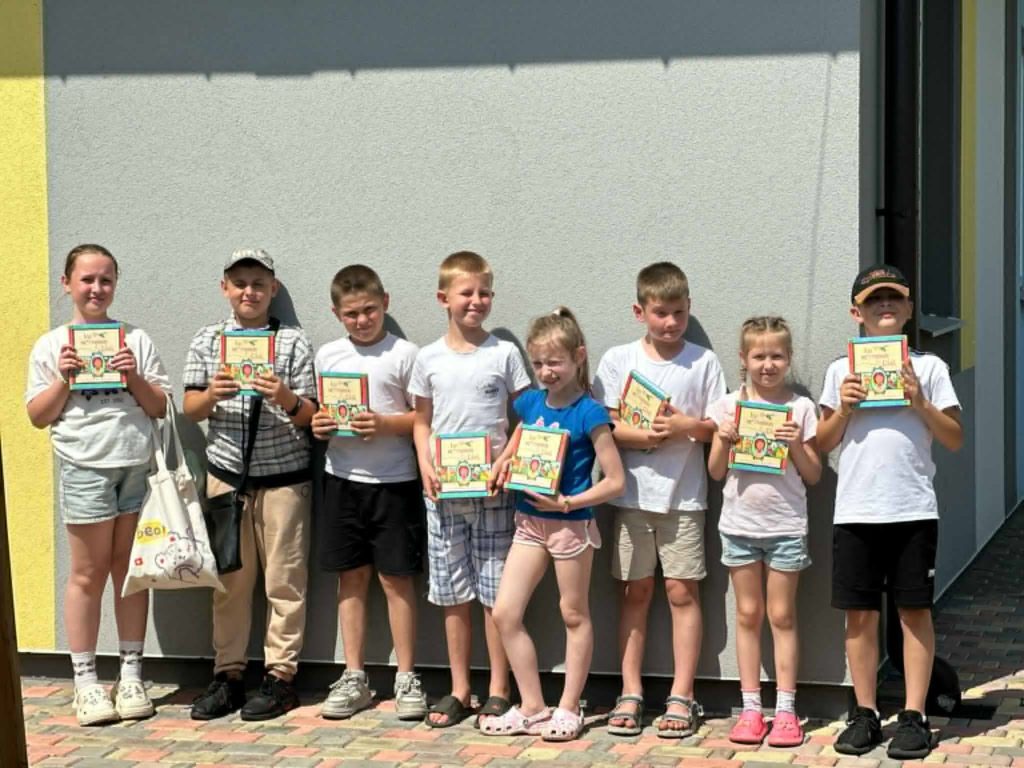 Nine children standing outside in a row against a light-coloured building wall, each proudly holding a copy of the Jesus Storybook Bible, participating in a summer camp on a sunny day.