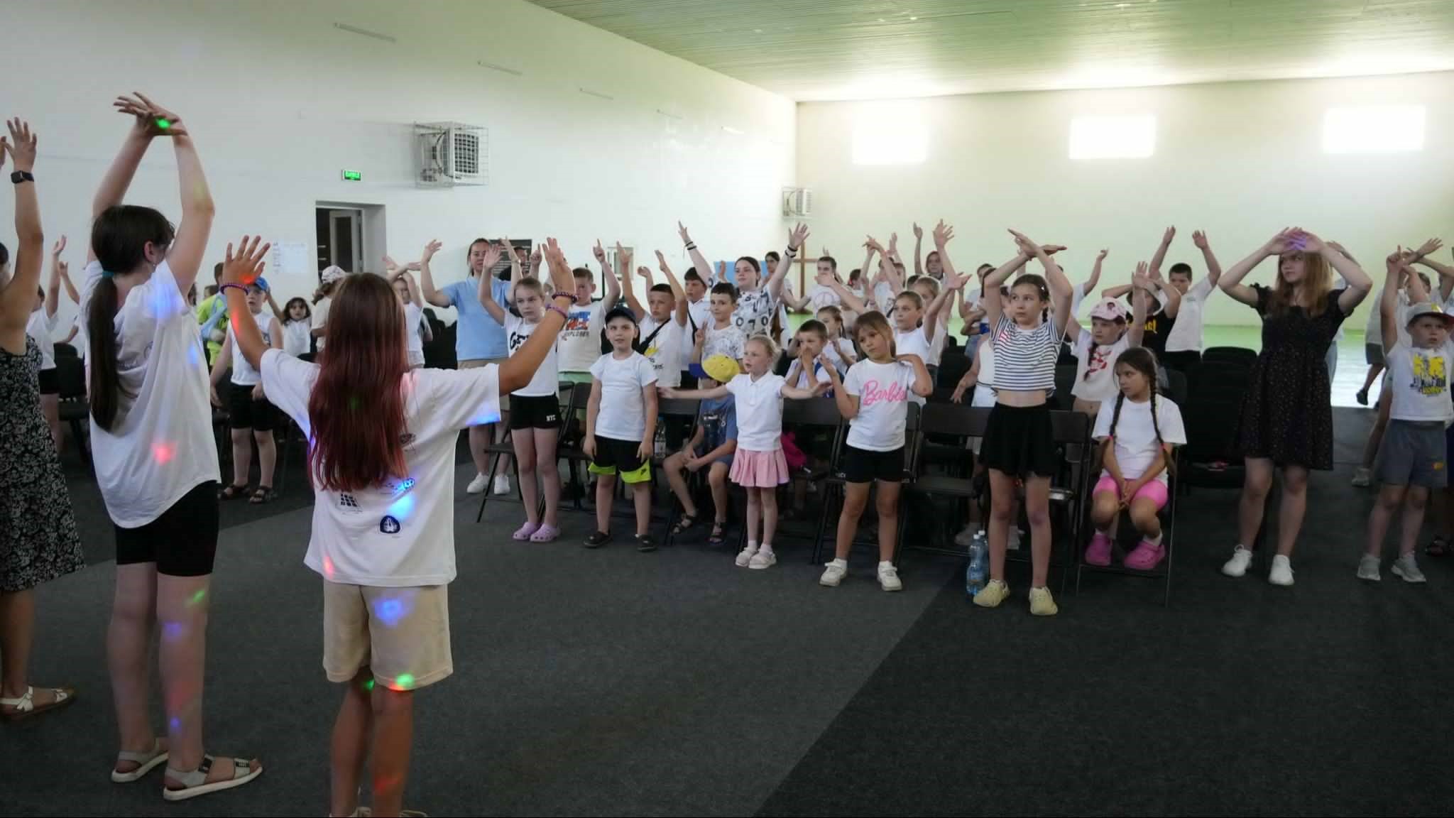 Large group of children and a few adults inside a brightly lit room, enthusiastically participating in an interactive activity with raised arms, possibly singing or dancing during a summer camp