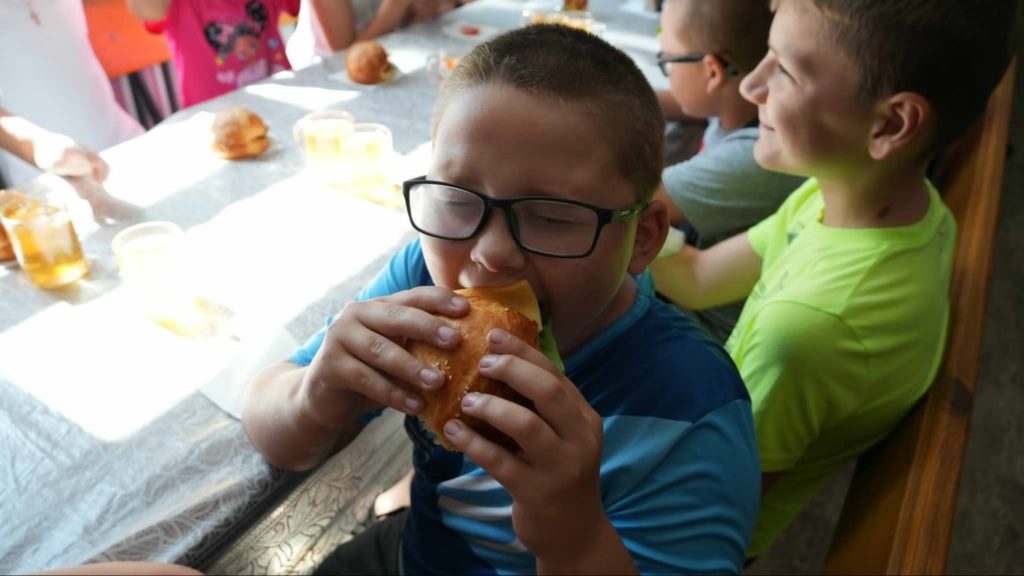 Young boy wearing glasses eagerly taking a big bite of a sandwich at a table surrounded by other children, all enjoying a meal together in a brightly lit dining setting.