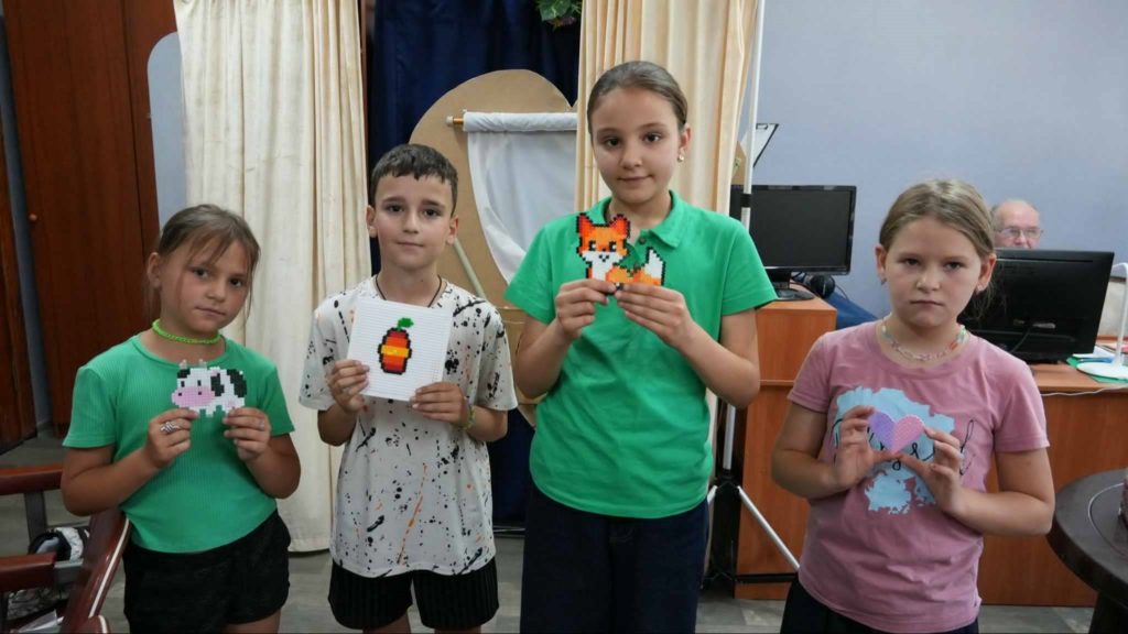 Four children standing indoors holding up pixel art crafts they created, including images of a cow, fruit, a fox, and a heart, proudly displaying their artwork at the summer camp