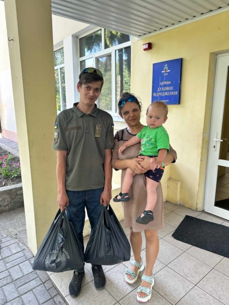 A man and woman stand outside a building holding a toddler and two large bags of aid.