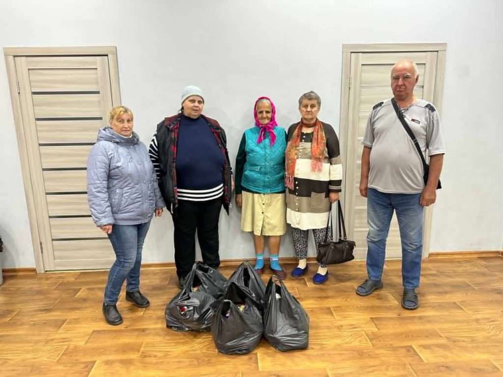 Five adults, including elderly women and men, standing indoors in a room with wooden flooring, posing behind several black plastic bags filled with food or supplies, representing a distribution effort for refugees or those in need.