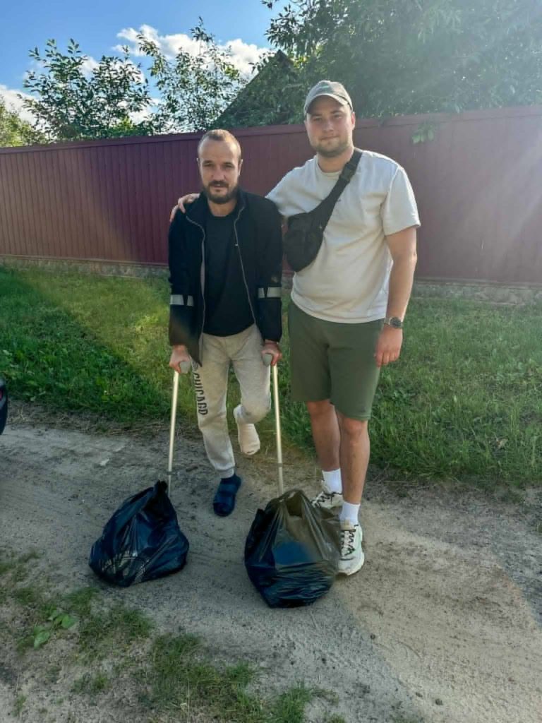 Two men standing outdoors on a dirt path next to a red fence, one using crutches, with black plastic bags filled with food or supplies at their feet, symbolizing humanitarian aid and support.