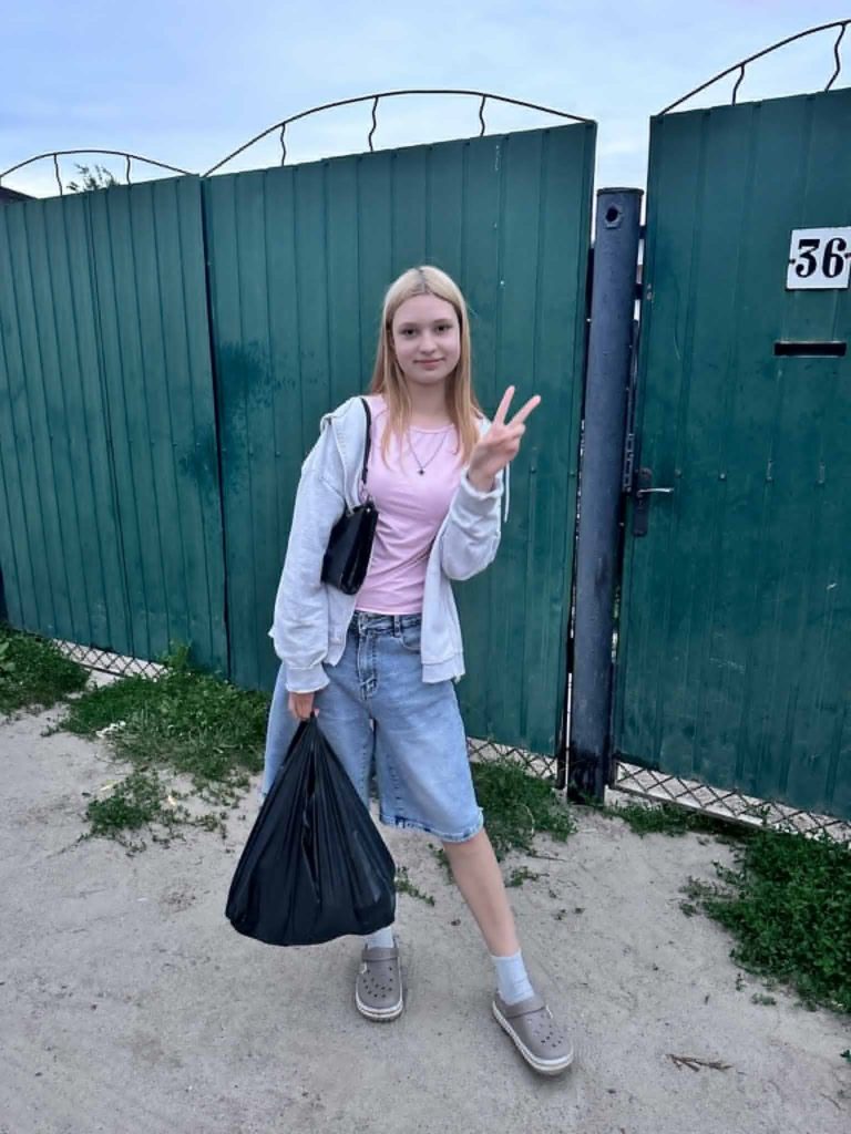 Teenage girl standing outside in front of a green metal gate, holding a black plastic bag filled with supplies in one hand and making a peace sign with the other, symbolizing aid distribution.