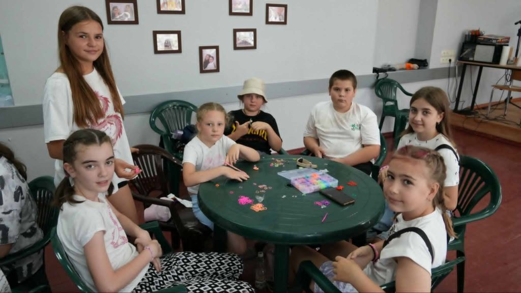 Group of children sitting around a green plastic table indoors, engaging in a craft activity with colourful beads and rubber bands, smiling and interacting in a relaxed, creative environment.