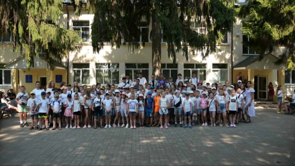 Children and adult leaders gathered outside the Centre under tall trees, posing for a group photo during the summer camp, with many wearing white shirts, hats, and smiles.