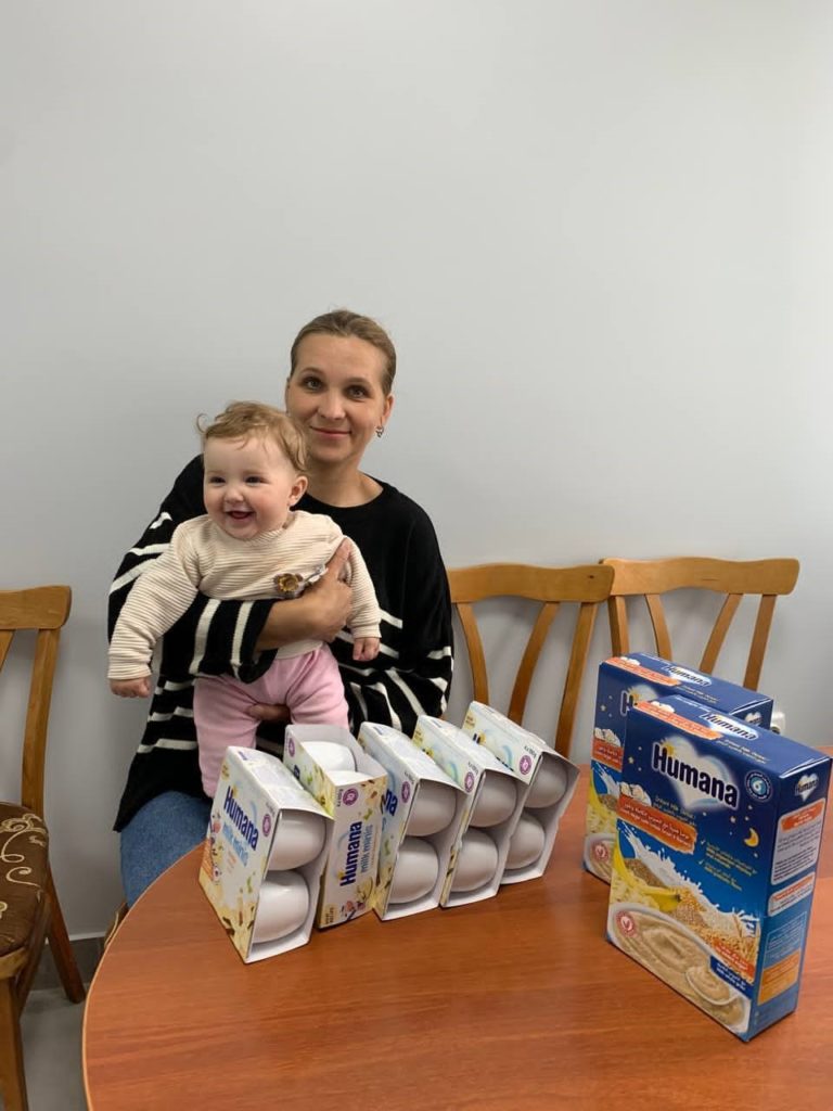 Smiling mother holding a happy baby while sitting at a table with several boxes of Humana baby formula and cereal, representing aid or support provided for mothers with young children.