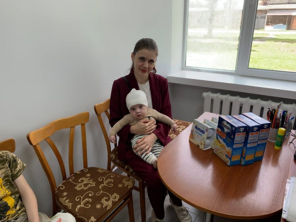 Young mother sitting by a window holding her baby on her lap, with boxes of Humana baby formula and cereal placed on the table beside them, representing assistance provided to mothers and infants.
