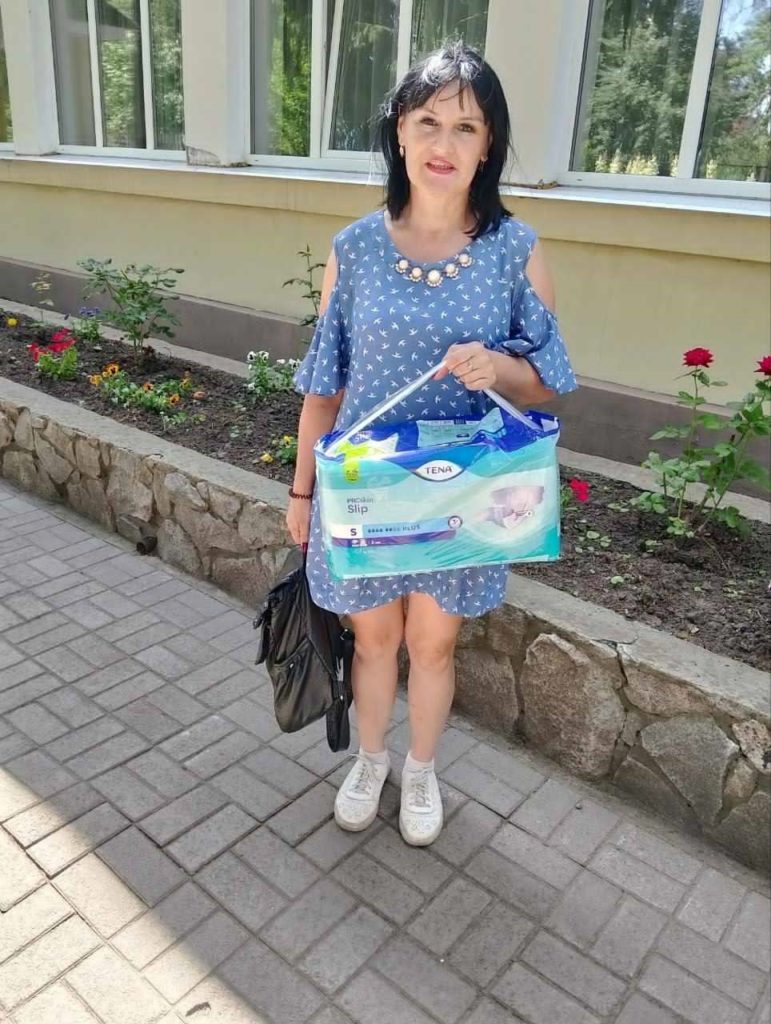 A woman in a blue dress with white patterns stands on a paved walkway holding a large pack of TENA incontinence products, with a black handbag over her arm, in front of a building with flowerbeds and windows.