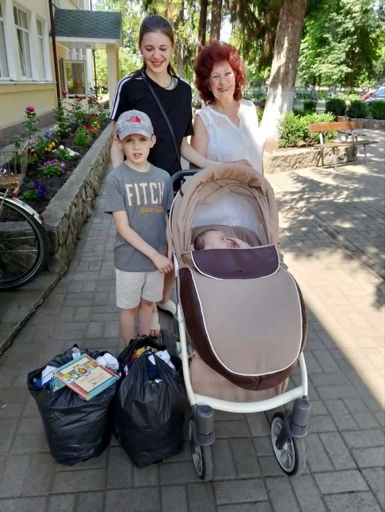 A smiling woman, a teenager, and two young children stand on a paved path with a baby in a pushchair and black bags of aid beside them, in front of a building with a flower bed.