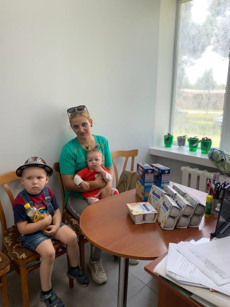 A mother sits indoors holding a baby while a young boy sits beside her, with various food items and baby formula stacked on a wooden table near a window