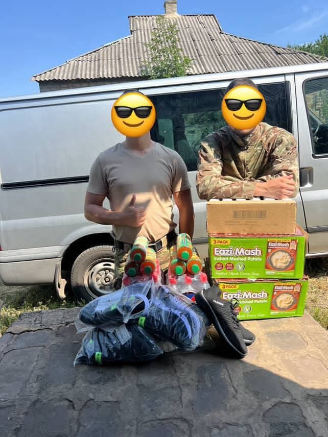 Two soldiers with sunglasses emoji over their faces standing beside bags of aid, including food and shoes, in front of a van.