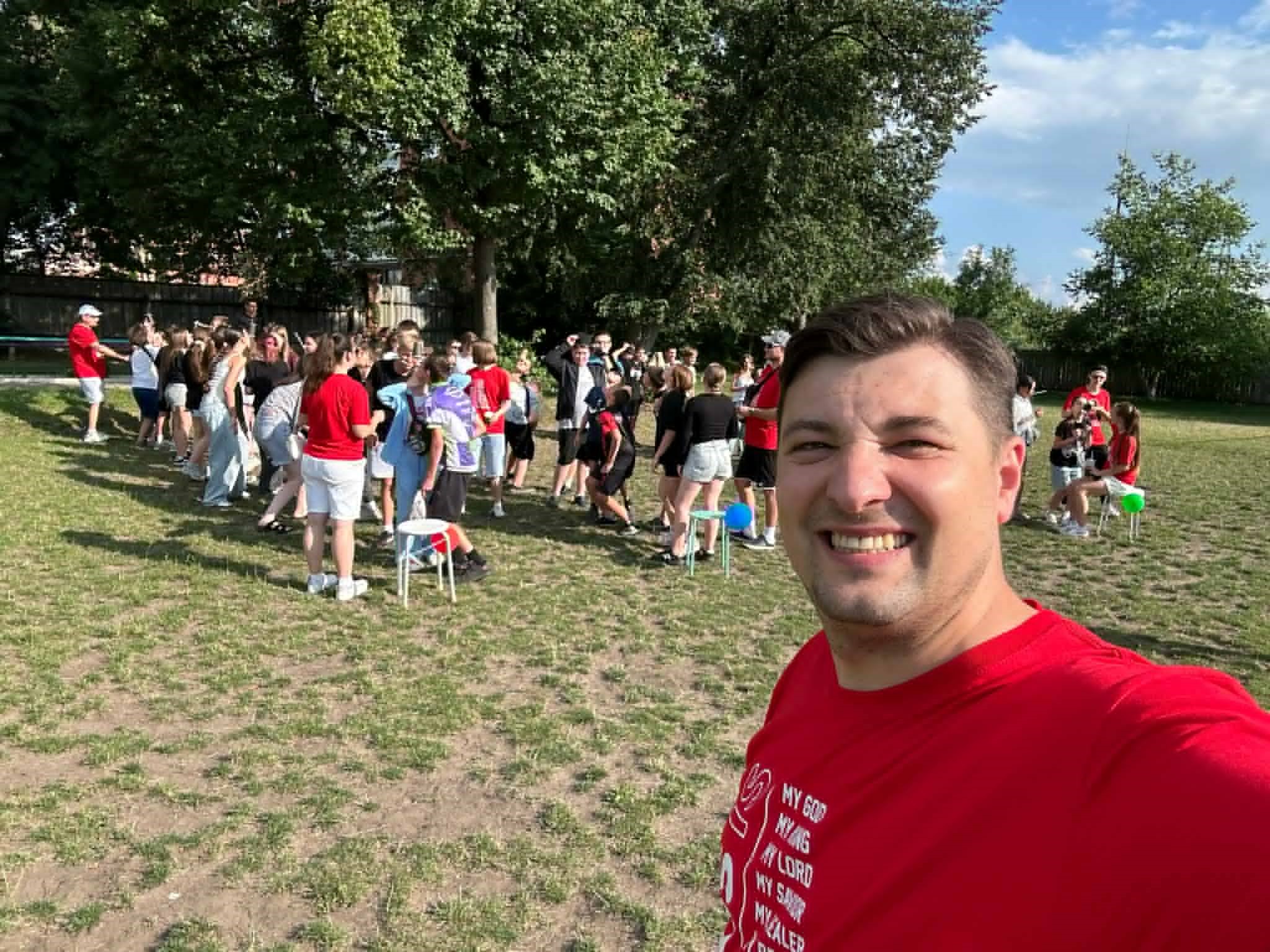 Selfie of a smiling Bohdan in the foreground with a large group of teenagers gathered on a grassy field in the background, participating in outdoor camp activities.