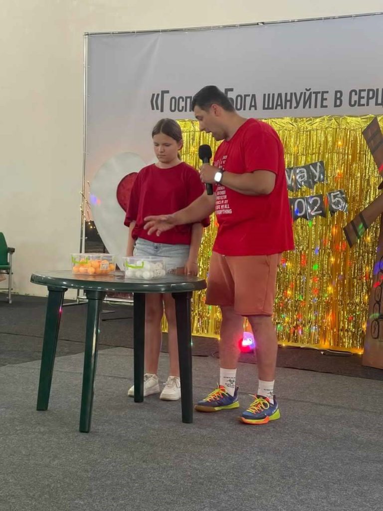 Bohdan and young girl in red shirts performing a game on stage with props on a table, in front of a decorative gold backdrop at a youth camp event.