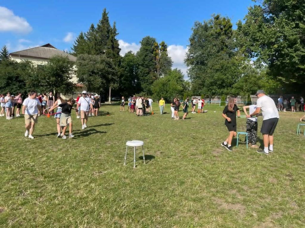 Multiple groups of teens and camp leaders engaged in outdoor relay games on a grassy field during a sunny day at youth camp.