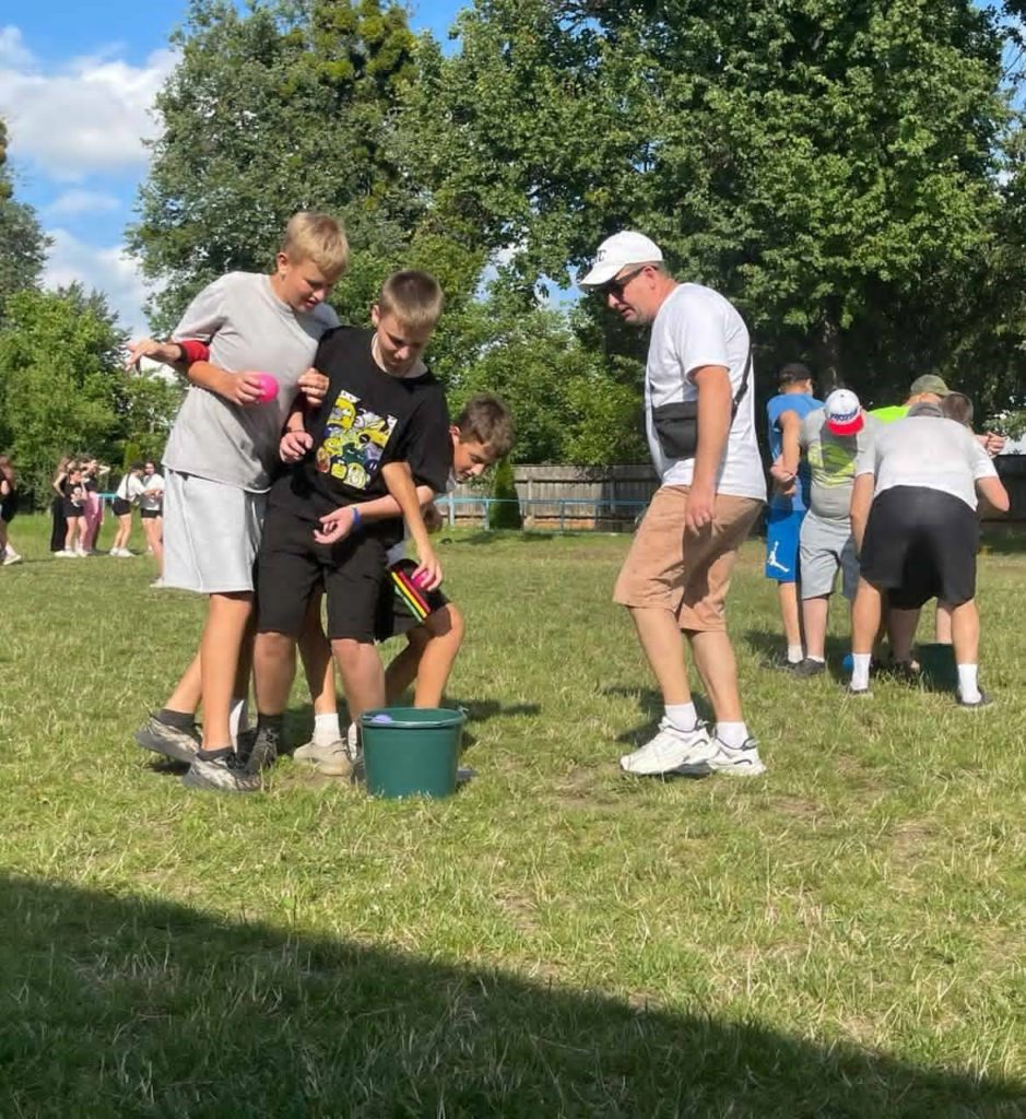 Boys in athletic wear participating in a team game involving buckets and coloured balls on a grassy field, supervised by camp leaders.