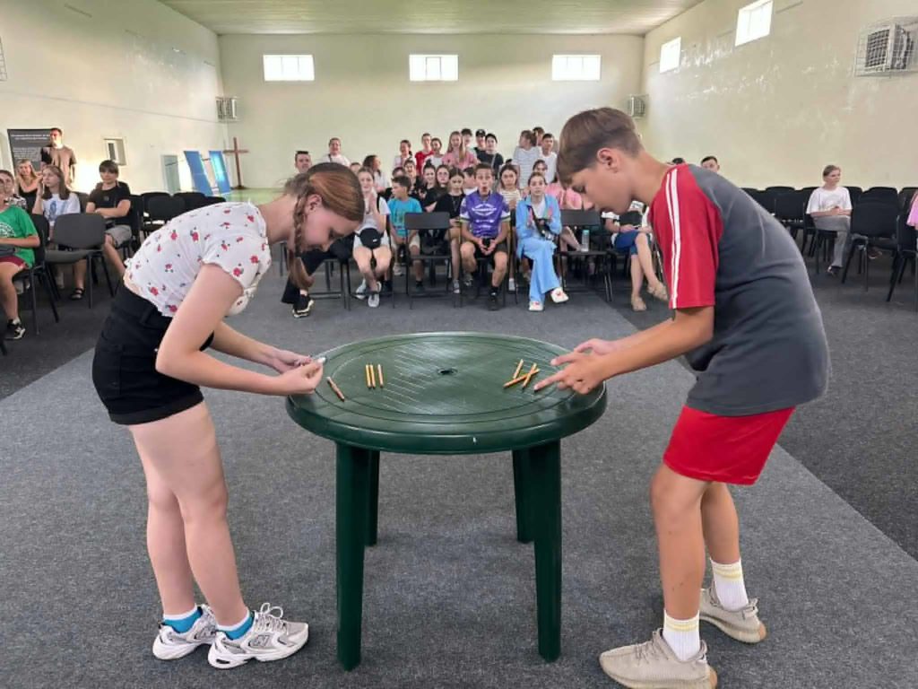Teen girl and boy concentrating on a tabletop game with pencils in front of an audience of other teenagers seated indoors.
