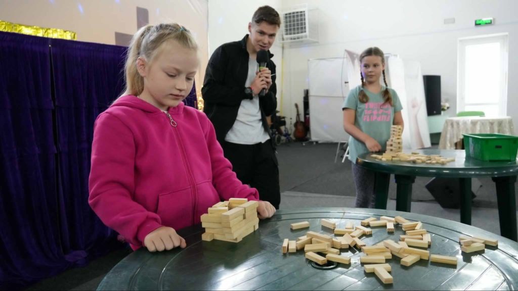Two young girls participating in a stacking game using wooden blocks at a summer camp, with a man holding a microphone observing and encouraging them in a brightly lit indoor setting.