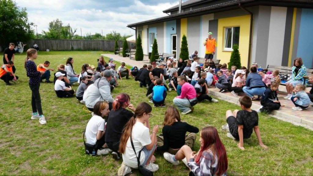 Large group of children gathered outdoors on the grass in front of a colorful building, participating in a summer camp activity led by an adult in a bright orange shirt, with some children sitting in circles and others watching.