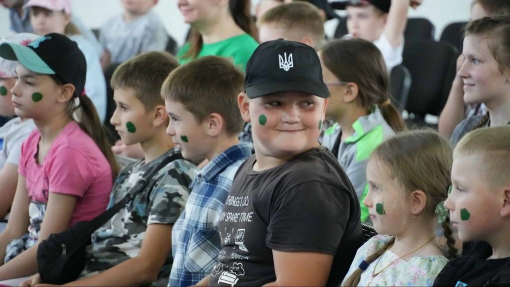 Children sitting closely together in a row, smiling and engaged in a group activity, each with a green dot painted on their cheek, during a summer camp indoor event.