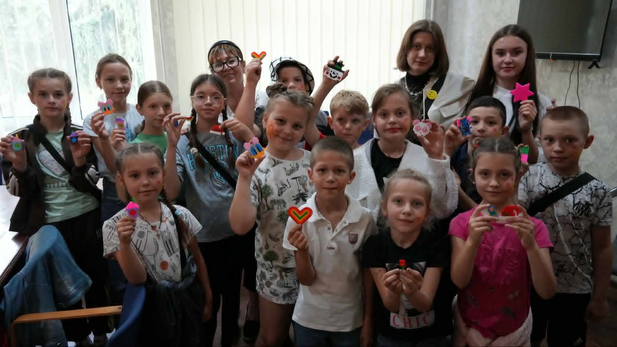 Group of smiling children standing indoors, proudly holding up colorful craft creations made from beads and other materials, with some having painted faces, during a summer camp activity.
