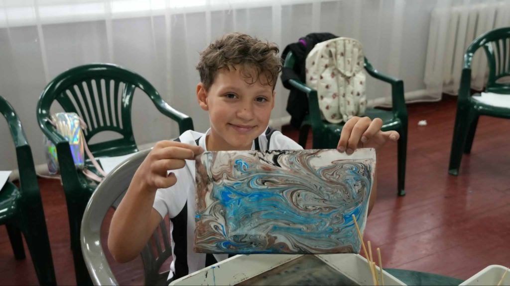 Smiling child sitting indoors holding up a piece of marbled artwork with swirls of blue, brown, and white, surrounded by plastic chairs and art supplies during a summer camp activity.