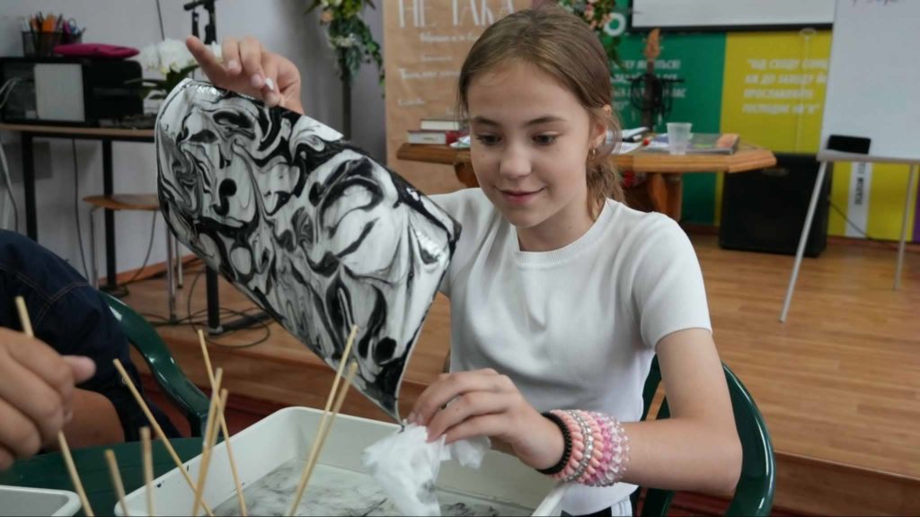 Young girl engaged in a marbling art activity, carefully lifting a black and white swirled design from a water tray, with art supplies and classroom materials visible in the background during a summer camp session.