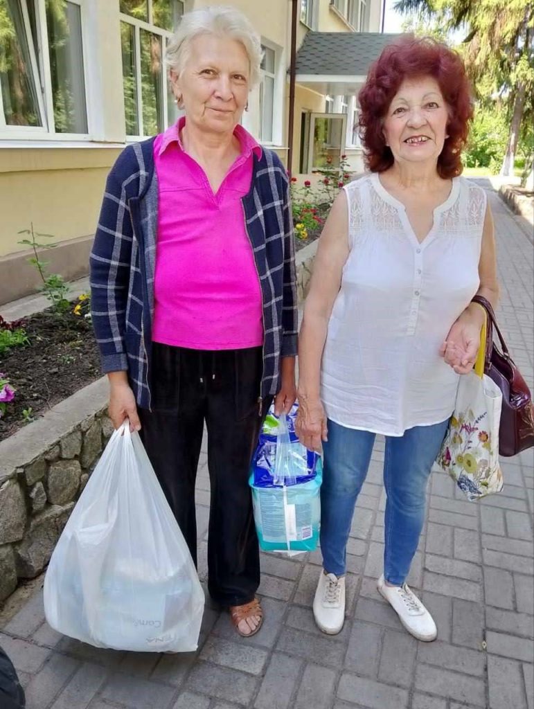 Two older women standing outside holding bags of aid, smiling in front of a building with flowerbeds