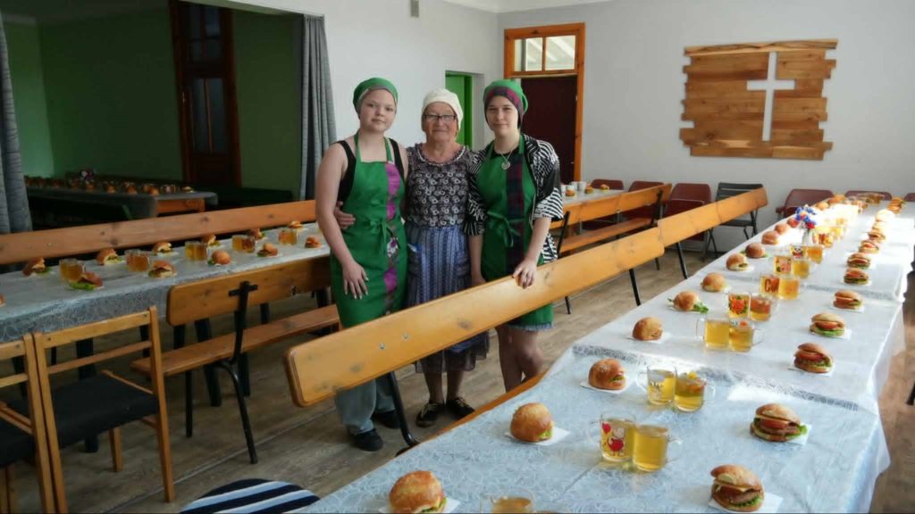 Three volunteers at the Centre, two in green aprons, standing in the dining hall with long tables set with burgers and drinks, and a wooden cross on the wall.