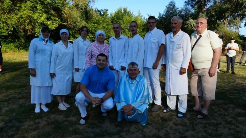 A group of men and women in white baptismal robes pose outdoors with Bohdan and Pastor Sergey in blue garments.