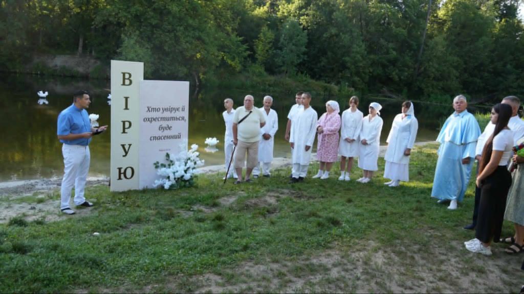 People in white baptismal robes stand by a riverbank with Bohdan speaking beside a large sign that reads “I believe” in Ukrainian.