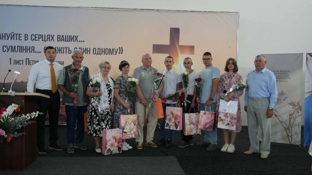Newly baptised individuals stand with flowers and gift bags in front of a church stage backdrop with a cross.