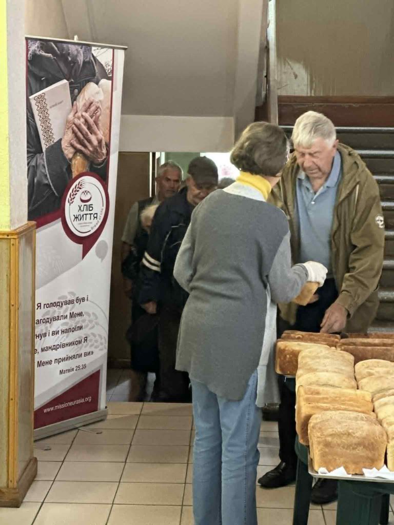 Volunteer handing fresh loaves of bread to elderly people in a hallway with a “Хліб Життя” (Bread of Life) banner.