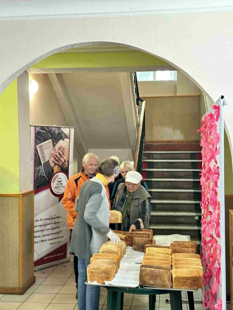 Group of elderly people receiving loaves of bread from a volunteer in an indoor setting.