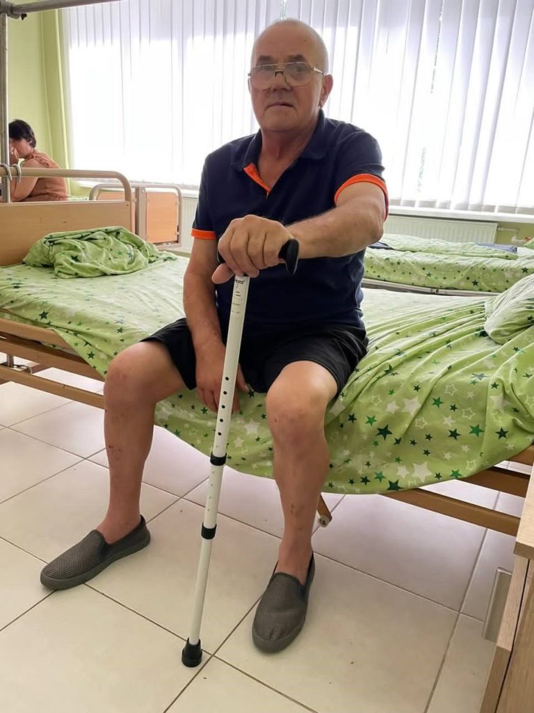 An older man wearing glasses sits on a bed in a hospital room holding a walking stick.