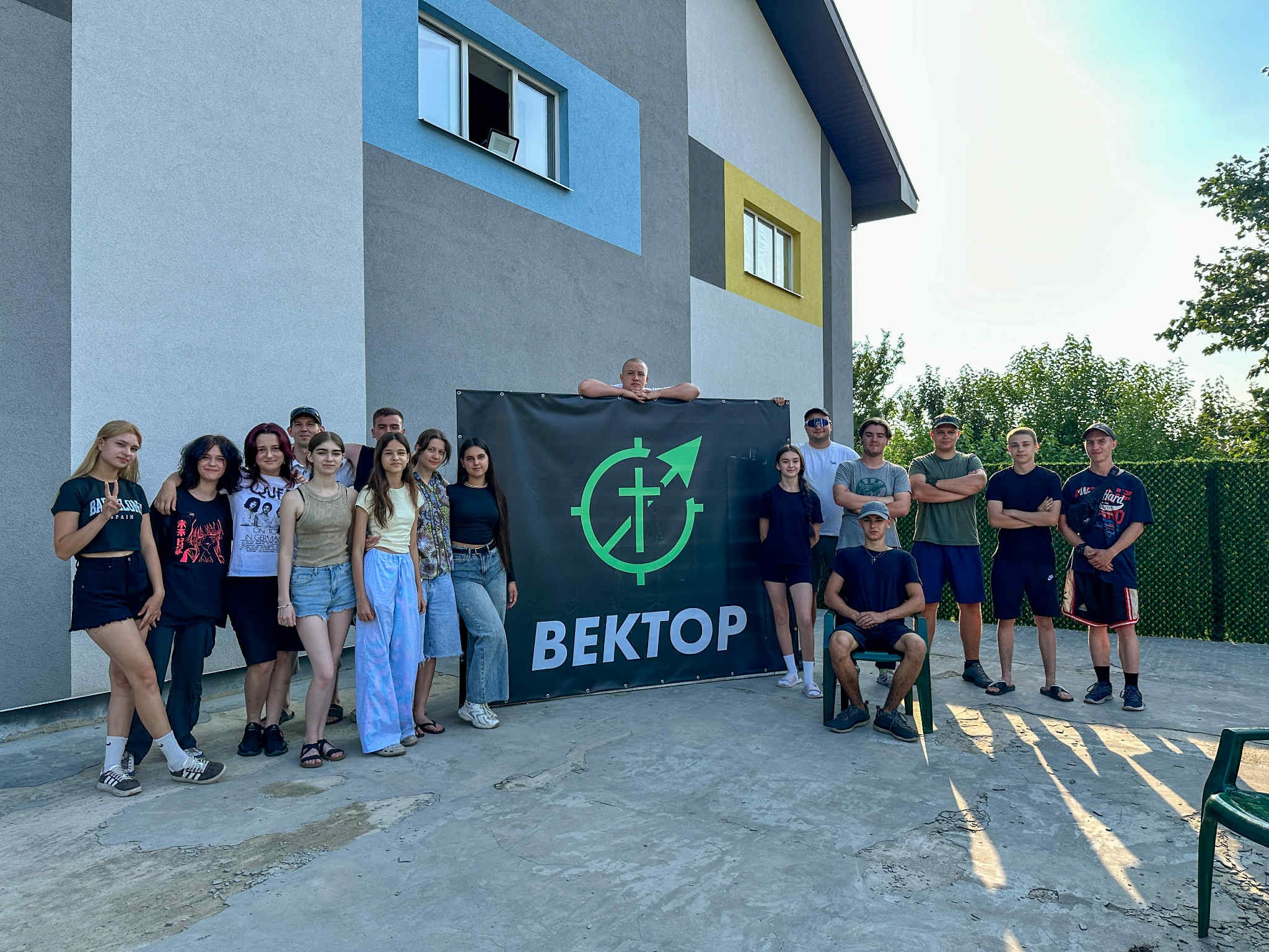 A group of teenagers and young adults stand and sit in front of a building around a large banner with the word Bektop on it.