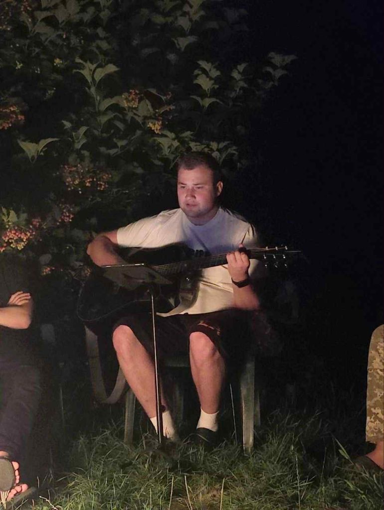 A young man sits on a plastic chair playing an acoustic guitar under dim light, with foliage in the background.
