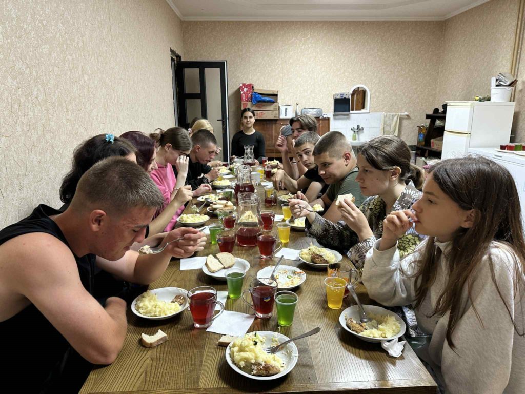 Several teenagers sit around a wooden table eating a meal together, with plates of mashed potatoes, bread, and drinks.