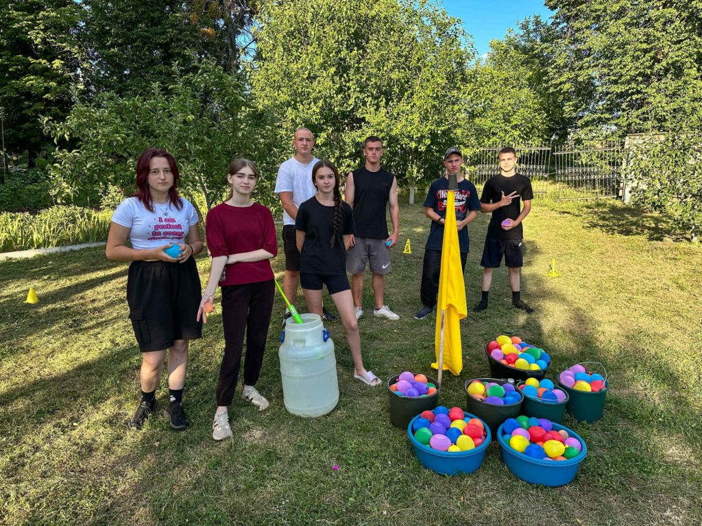 A group of young people stand outside on grass beside buckets filled with colourful water balloons, preparing for a game.