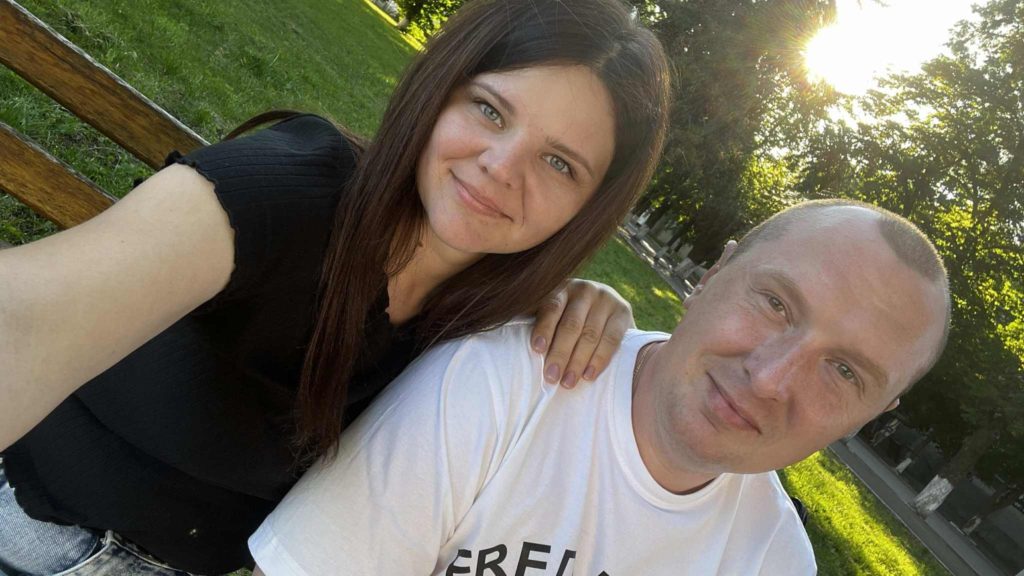 A smiling young couple sitting on a park bench with the sun setting behind them, the woman’s hand resting on the man’s shoulder.