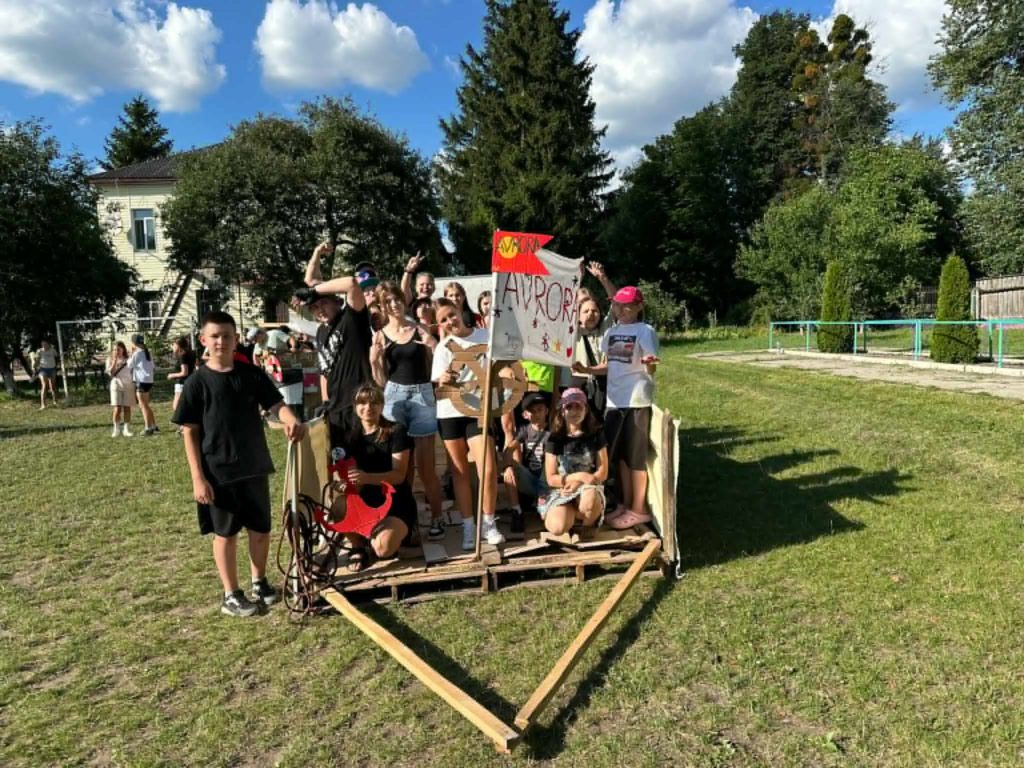 Group of teenagers posing together with a hand-made banner and wooden cart structure during a camp activity.