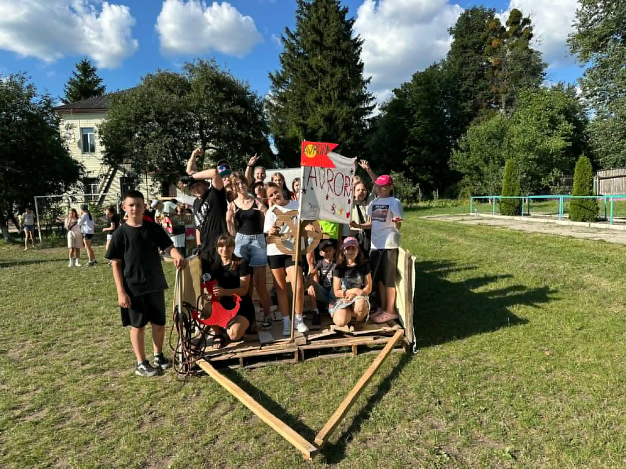Group of teenagers posing together with a hand-made banner and wooden cart structure during a camp activity.