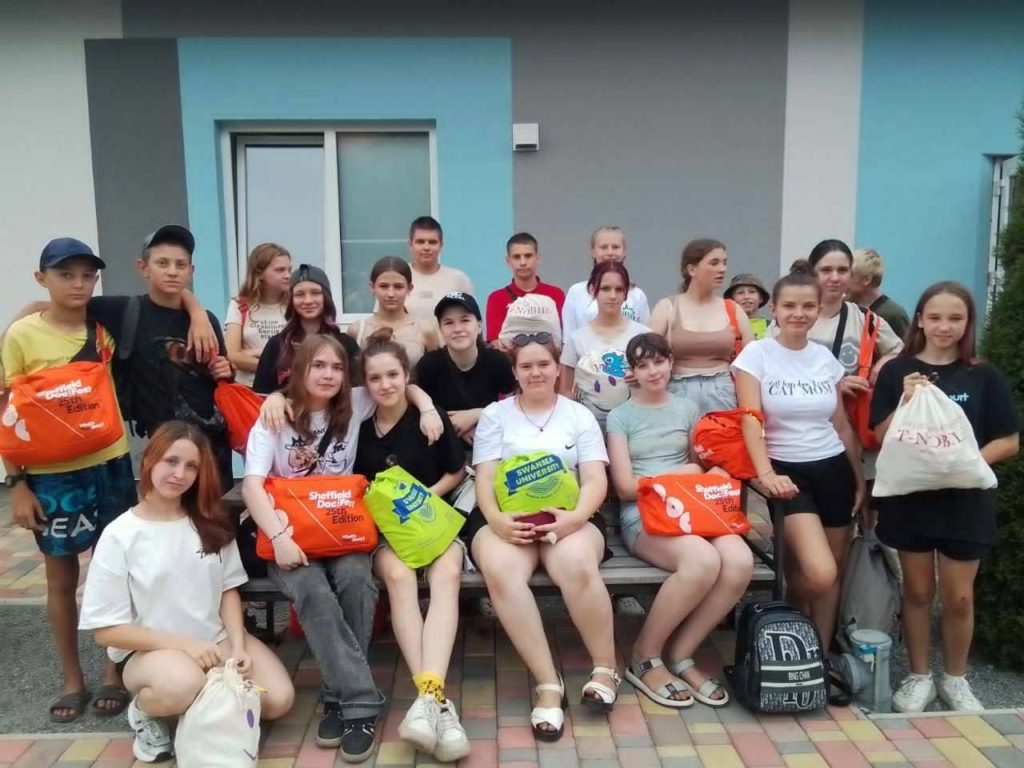 Group photo of teens holding colorful bags, standing together in front of a building during summer camp.
