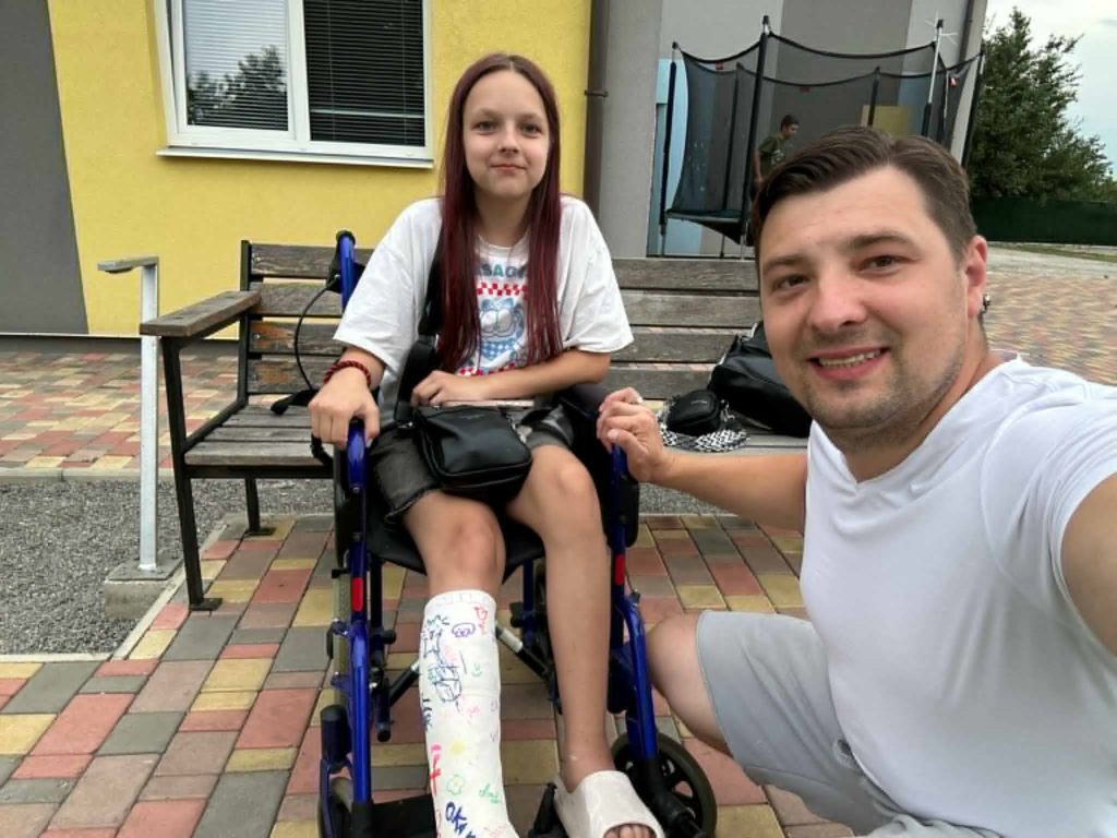 Smiling teen girl in a wheelchair with a signed leg cast, posing with a Bohdan in a white outfit outside a building.