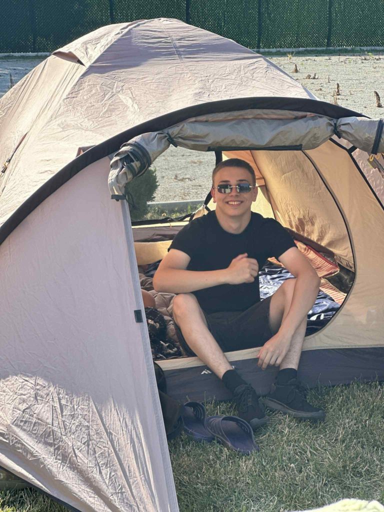 Young man sitting inside an open tent pitched on grass, smiling and wearing sunglasses.