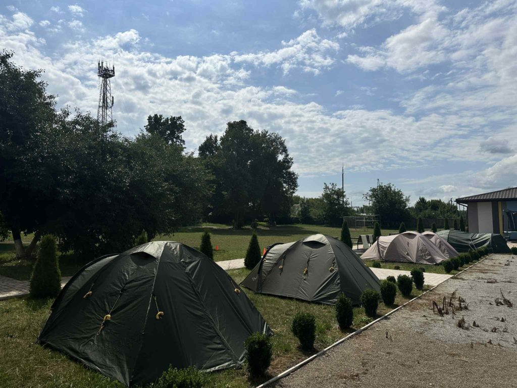 Row of pitched tents on grass near the Centre building and trees, under a partly cloudy sky.