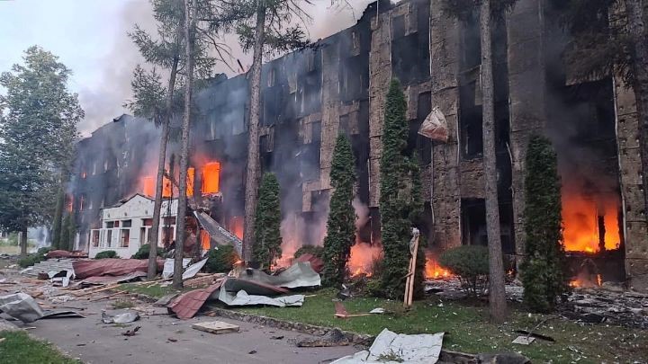 University building engulfed in flames after a bombing, with smoke rising and debris scattered in front.